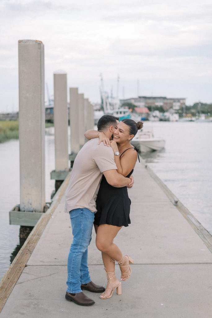 Man kissing his fiancé wearing a black dress on a dock at Shem Creek South Carolina with boats in the background  