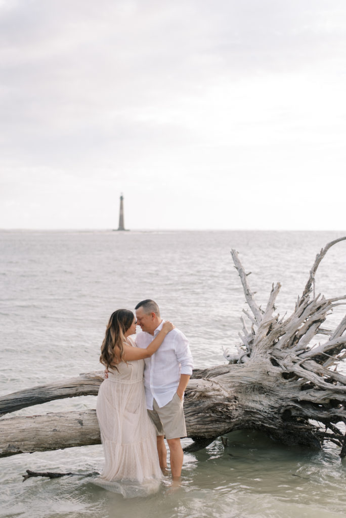 Husband and wife sitting on driftwood in the water with Morris Island Lighthouse in the background on Folly Beach South Carolina wearing khaki and white and an ivory lace dress