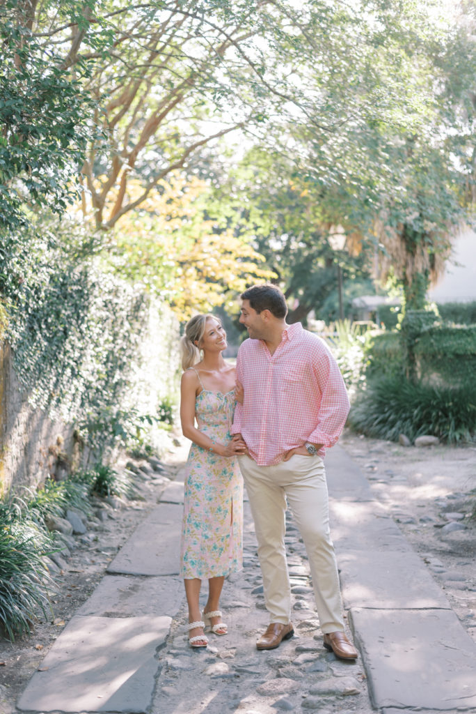 Engaged couple wearing flower sundress and pink shirt standing together in Longitude Lane with brick and ivy in Charleston