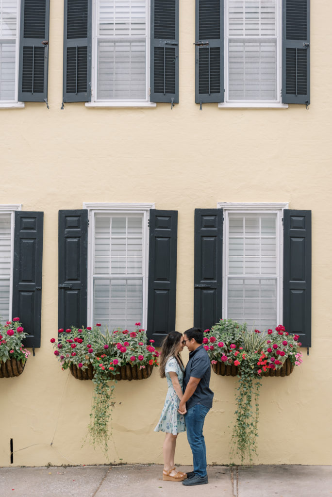 Engaged couple standing together in front of yellow building with pink flower boxes on Tradd St in Charleston South Carolina