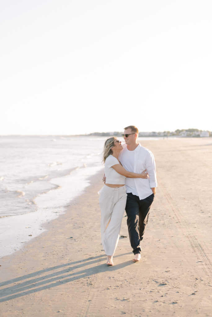Engaged couple hugging and walking on Isle of Palms beach at sunset wearing sunglasses