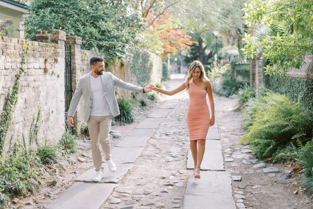 Engaged couple walking through an alley on cobblestone with brick wall and ivy in downtown Charleston