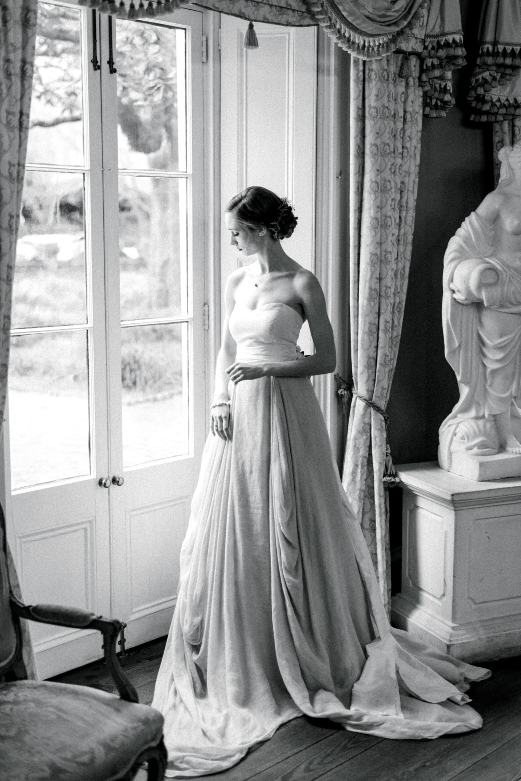The black and white image shows a beautiful bride standing inside the William Aiken House. She is wearing a strapless corset top and a blush-colored wedding skirt that flows down to the floor. The corset top is fitted to her body, accentuating her curves, while the skirt has a soft and delicate texture, adding a touch of elegance to her appearance. The bride stands confidently, with her arms slightly lifted, and her face is turned towards the window, revealing a peaceful and content expression. The black and white effect of the photo adds a classic and timeless feel to the image, highlighting the bride's beauty and poise.