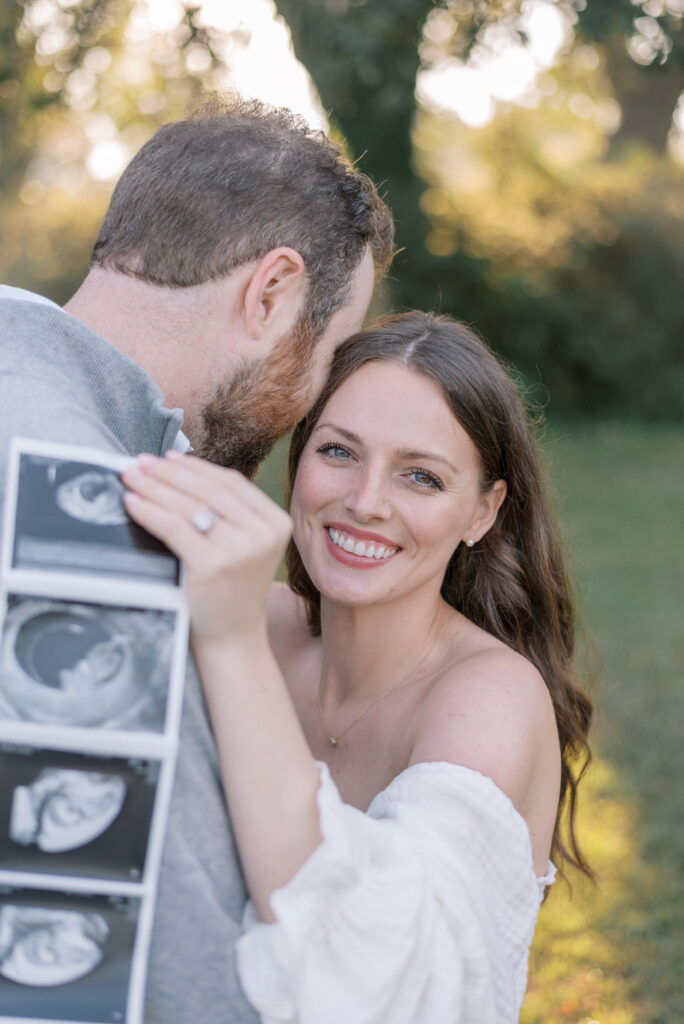 Woman smiling at the camera hugging her husband and holding her ultrasound photos