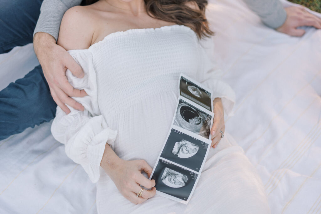 Pregnant woman in a white dress laying on a blanket with ultrasound photos
