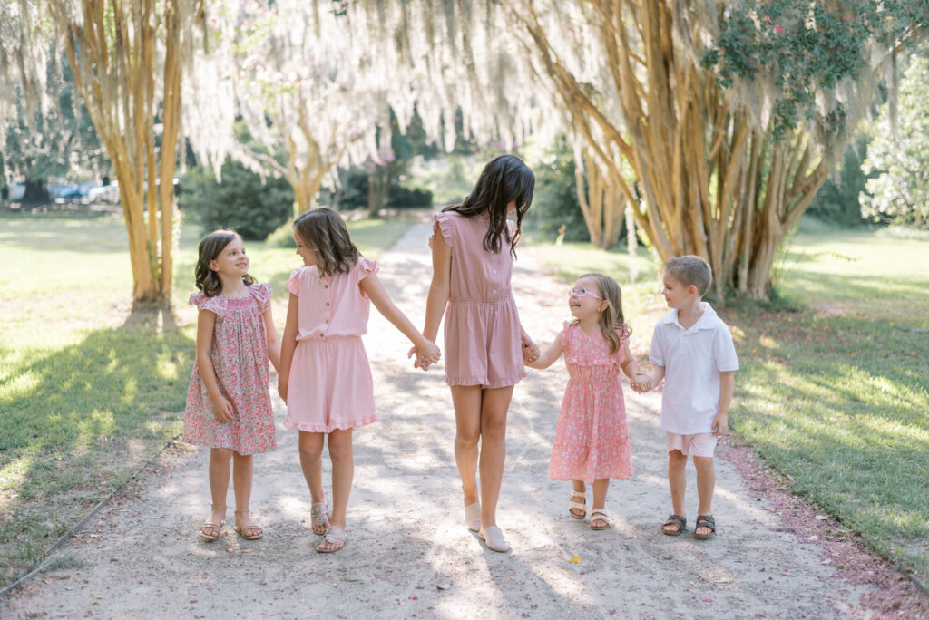 Children wearing pink and holding hands walking through Hampton Park under Crepe Myrtle trees and Spanish Moss for family portraits