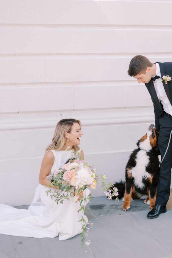 A bride and groom on their wedding day, the bride is kneeling down and laughing while holding her bouquet as the groom attempts to get their dog to behave.