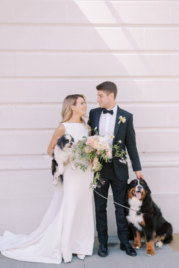 A bride and groom smiling at each other on their wedding day. The bride holding their small fluffy dog and their bernese mountain dog sitting politely beside them