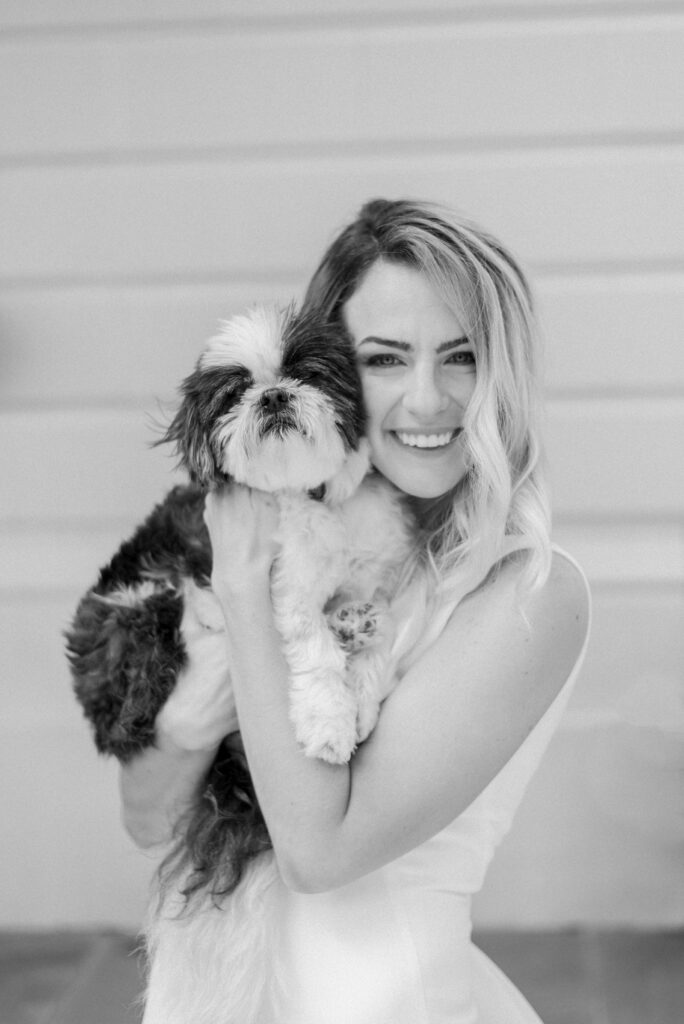 A black and white photograph of a bride holding her dog close and smiling at the camera. The bride is wearing her wedding dress and the dog is small and fluffy.