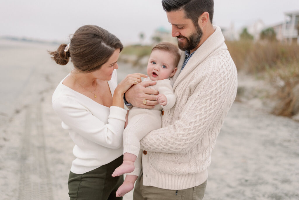 Happy family of three on the beach at sunrise, holding their adorable baby boy in a cute ivory sweater and pants outfit.