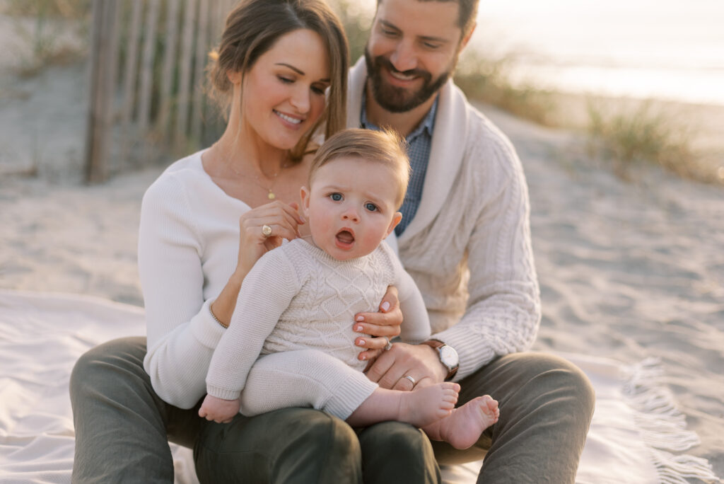 A family is depicted in a photograph sitting on a white blanket on the beach at sunrise. The mother and father are seated with their baby, and the baby is looking towards the viewer with an adorable expression. The parents are smiling and looking lovingly at their child. The sand dunes provide a beautiful and natural backdrop, with the waves gently lapping against the shore. The golden hues of the sunrise cast a warm glow on the family, creating a serene and peaceful atmosphere. The image captures a precious moment of love and tenderness between a family, as they enjoy a beautiful morning on the beach together.