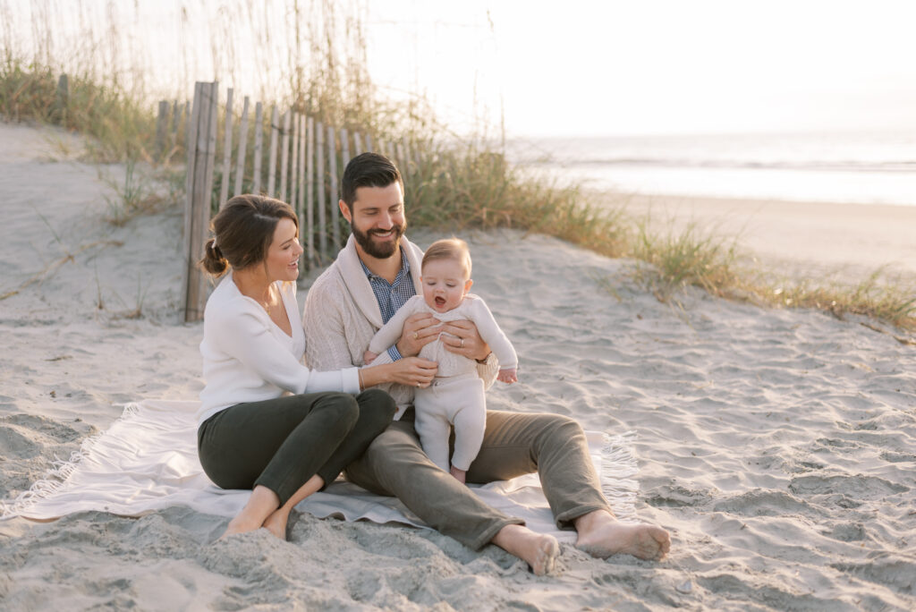 A family is captured in a photograph sitting on a white blanket on the beach at sunrise. The mother and father are seated with their baby, and all three are laughing joyfully. The sand dunes provide a beautiful and natural backdrop, creating a serene and tranquil atmosphere. The golden hues of the sunrise cast a warm glow on the family, emphasizing the beauty and happiness of the moment. The image portrays a family enjoying a beautiful morning on the beach together, creating memories and sharing love and laughter in the midst of a beautiful natural setting.