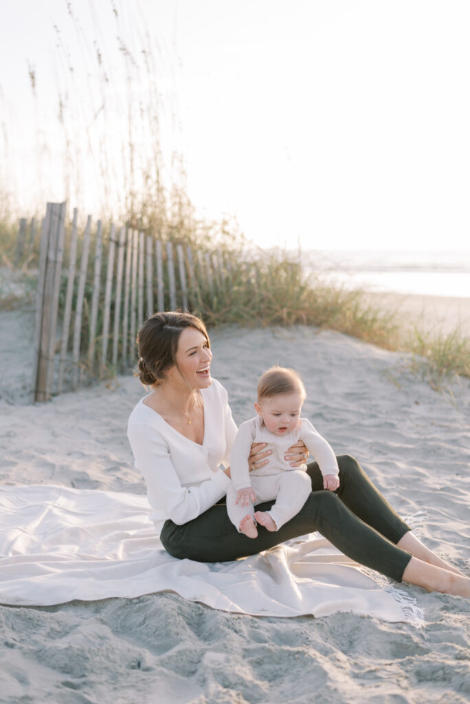 An image shows a mother sitting on a white blanket on the beach at sunrise, holding her baby and laughing joyfully. The baby is sitting on the mother's knee, looking down with a content expression. The serene ocean is visible in the background, and the sand dunes provide a natural and tranquil backdrop. The warm and golden hues of the sunrise cast a beautiful light on the mother and child, creating a peaceful and happy atmosphere. The image captures a special moment of love and happiness between a mother and her child, as they enjoy a beautiful morning together on the beach.
