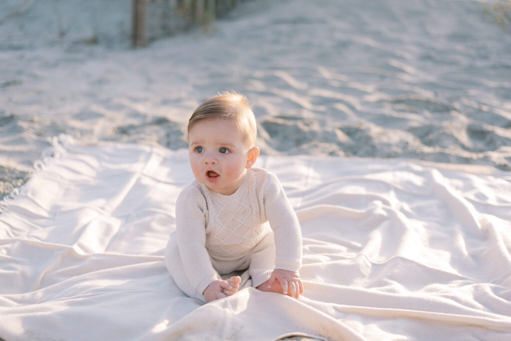 a baby sitting by himself on a white blanket on the beach at sunrise, gazing serenely forward towards the viewer. The tranquil ocean is not in view, but the soft sand and gentle sand dunes create a natural and peaceful background. The warm and golden hues of the sunrise cast a beautiful light on the baby, creating a serene and calm atmosphere. The image captures a precious moment of innocence and tranquility, as the baby sits in quiet contemplation on the beach, enjoying the serenity of the early morning