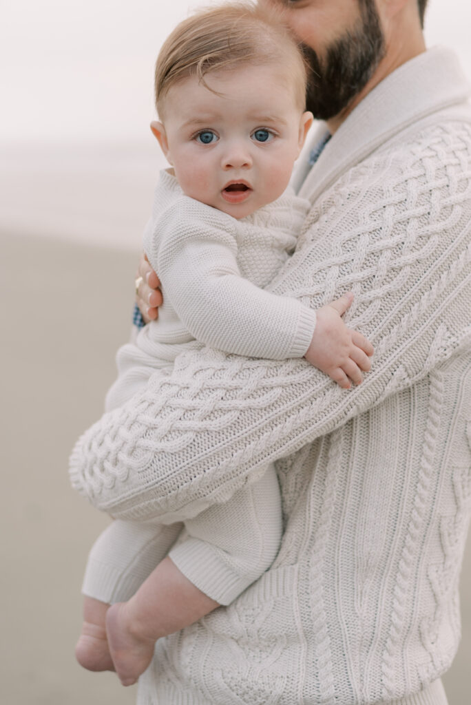 Close up of baby boy with blue eyes being held by his father on the beach during a sunrise family photo session.