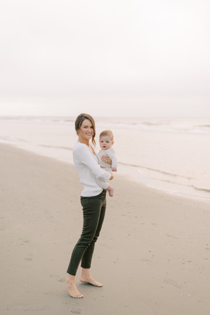Mother holding her baby on the beach at sunrise. The mother wears green pants and a white sweater while the baby is dressed in an ivory sweater outfit.