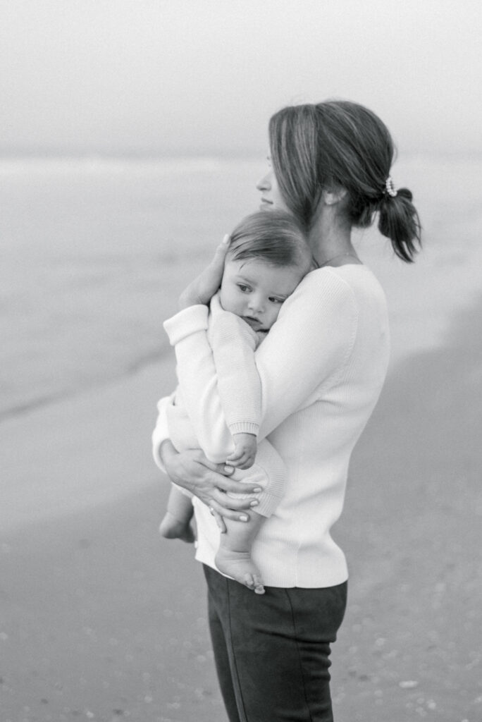 A mother is depicted in a black and white photograph, holding her child and gazing out at the ocean. The child is snuggled into the mother's shoulder, as if seeking comfort and security. The simplicity of the black and white image emphasizes the tenderness of the moment, as the mother and child share a quiet and peaceful moment together. The ocean provides a beautiful and serene backdrop, with the waves gently lapping against the shore. The image portrays a special bond between a mother and her child, as they share a moment of calm and tranquility in the midst of a vast and wondrous natural setting.
