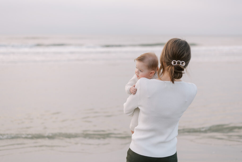 A mother is captured in a photograph holding her baby in front of the ocean. The mother is standing with her back to the camera, while the baby looks over her shoulder towards the viewer. The mother's hair is styled in a low bun, held in place with a pearl hair clip, giving her a classic and elegant appearance. The ocean creates a serene and tranquil background, with the waves gently crashing against the shore. The image portrays a tender moment between a mother and her child, as they admire the beauty of nature together.