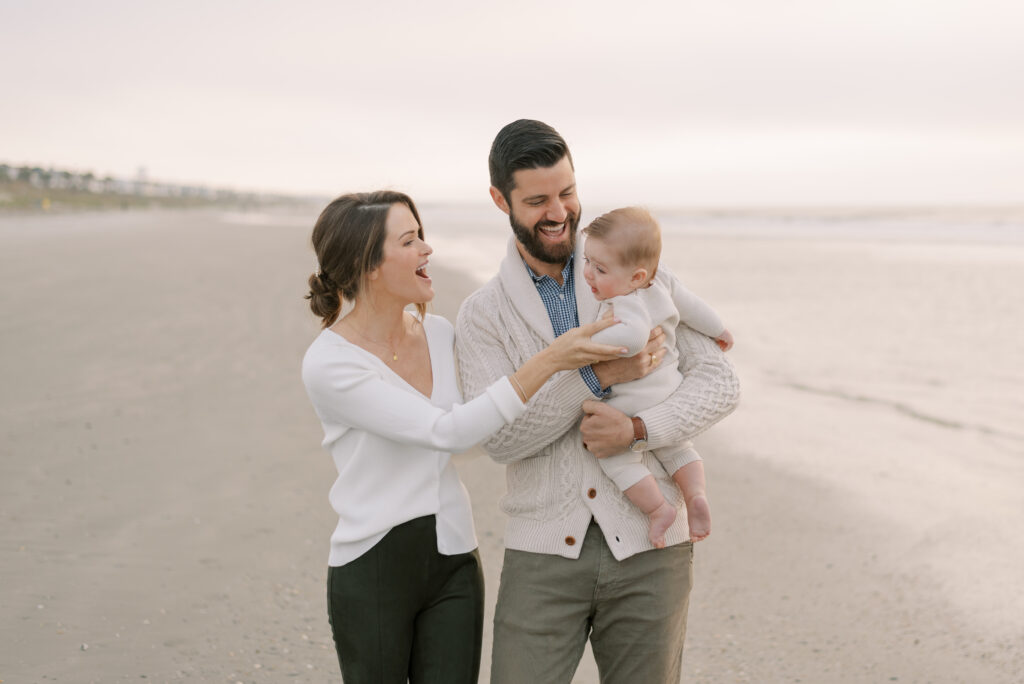 A family is depicted in a photograph walking on the beach during a sunrise photo session. The mother and father are holding their baby, and all three are laughing joyfully. The golden hues of the sunrise reflect beautifully on the ocean waves behind them, creating a serene and peaceful atmosphere. The parents are casually dressed in comfortable clothing, with bare feet in the sand. The image captures a moment of pure happiness and love, as the family enjoys a beautiful morning on the beach together.