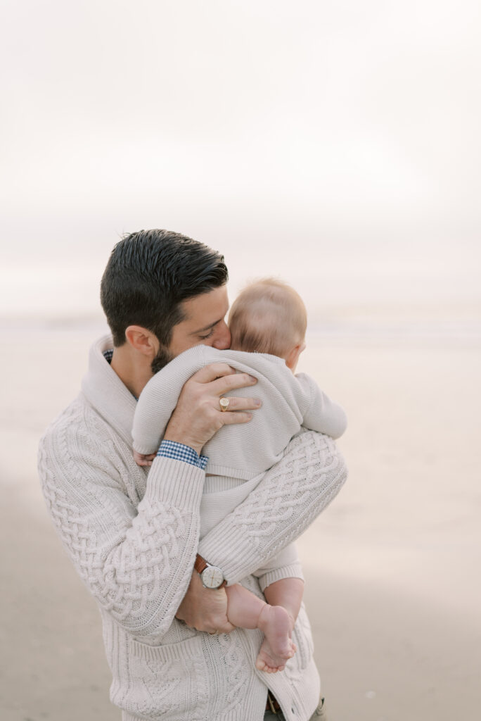 A father is depicted in a photograph holding his baby on the beach at sunrise. The father is giving the baby tender kisses, while the baby is snuggled into his shoulder. The golden hues of the sunrise cast a warm glow on the father and baby, creating a serene and peaceful atmosphere. The beach provides a beautiful and natural backdrop, with the waves gently lapping against the shore. The image captures a moment of pure love and tenderness between a father and his child, as they enjoy a beautiful morning on the beach together.