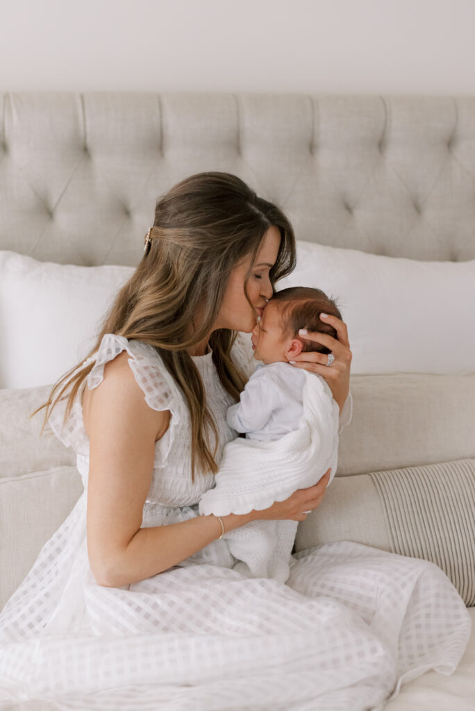 A tender moment captured in this image as a loving mother gazes at her precious newborn, cradling him gently in her arms. She wears a beautiful white dress, and her eyes are full of joy and tenderness as she plants a soft kiss on her baby's forehead. The intimate setting of the bedroom adds to the warmth and comfort of the scene. This image radiates pure love and maternal affection.