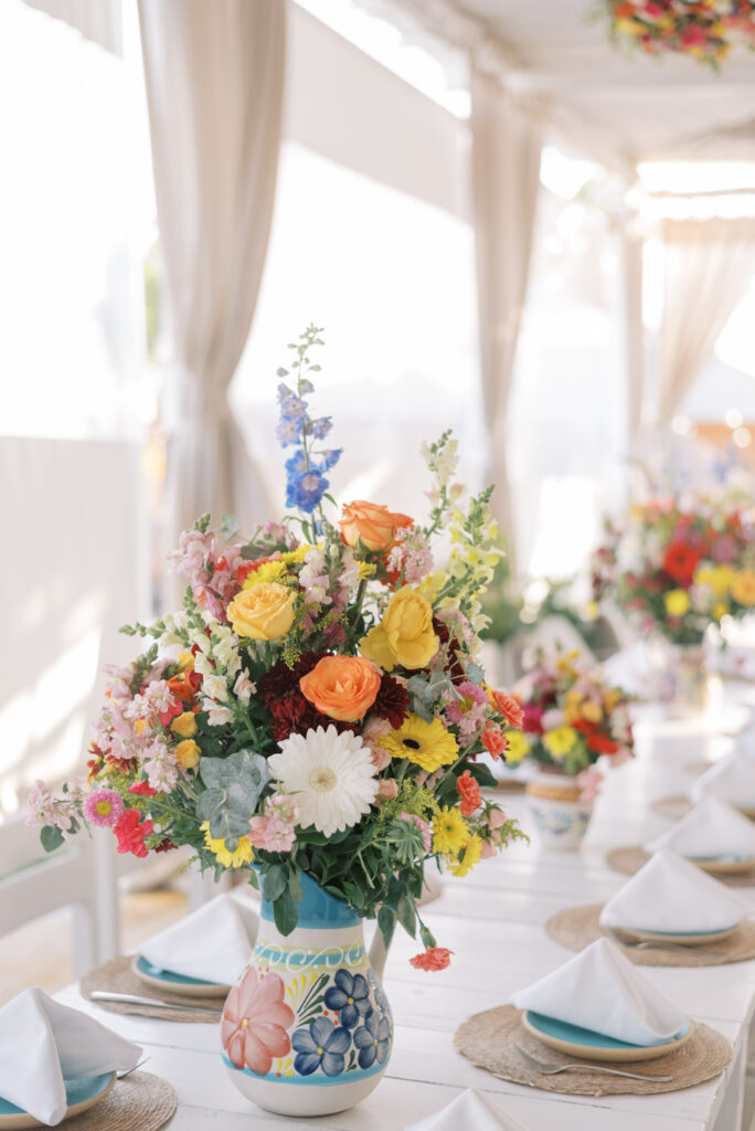 Bright and colorful floral centerpiece in a traditional Mexican pottery vase featuring vibrant orange, yellow, pink, white, blue, and green florals, beautifully complementing the stoneware dishes and creating a stunning table setting.