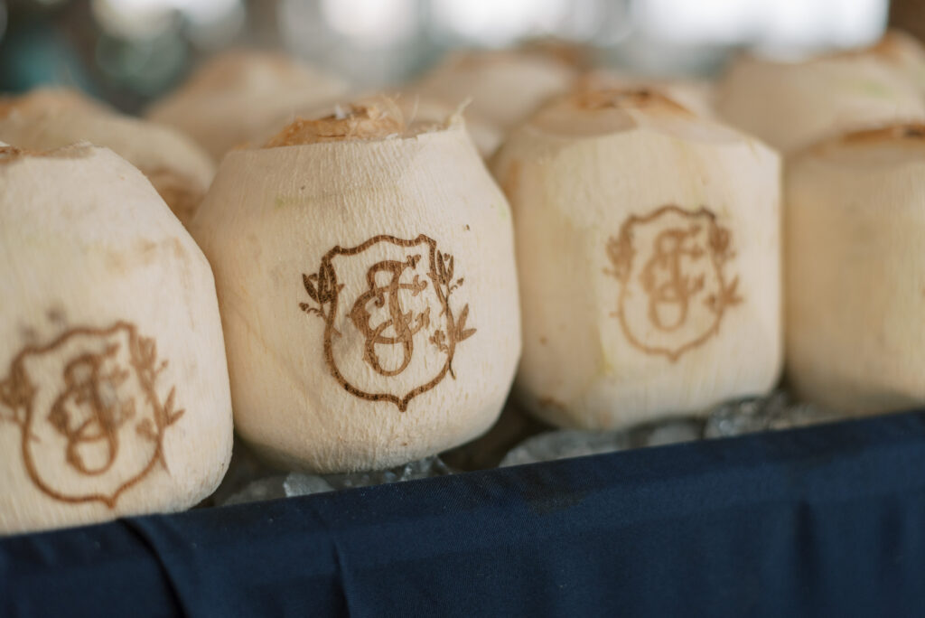Custom engraved coconut cups with a bride and groom crest design at a beachside rehearsal dinner in Akumal, Mexico. The cups are filled with a tropical drink and surrounded by colorful flowers and traditional Mexican pottery.