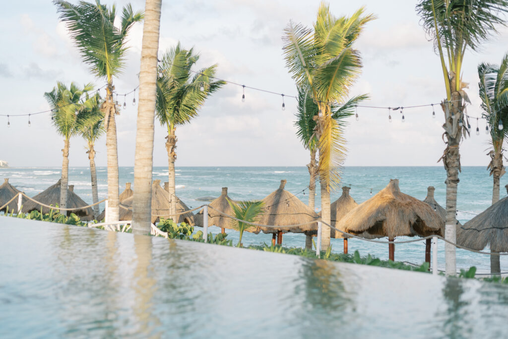 An infinity pool with a stunning ocean view, surrounded by lush palm trees and traditional thatched tiki huts. The pool is located in Akumal, Mexico, the perfect destination for a beach wedding.