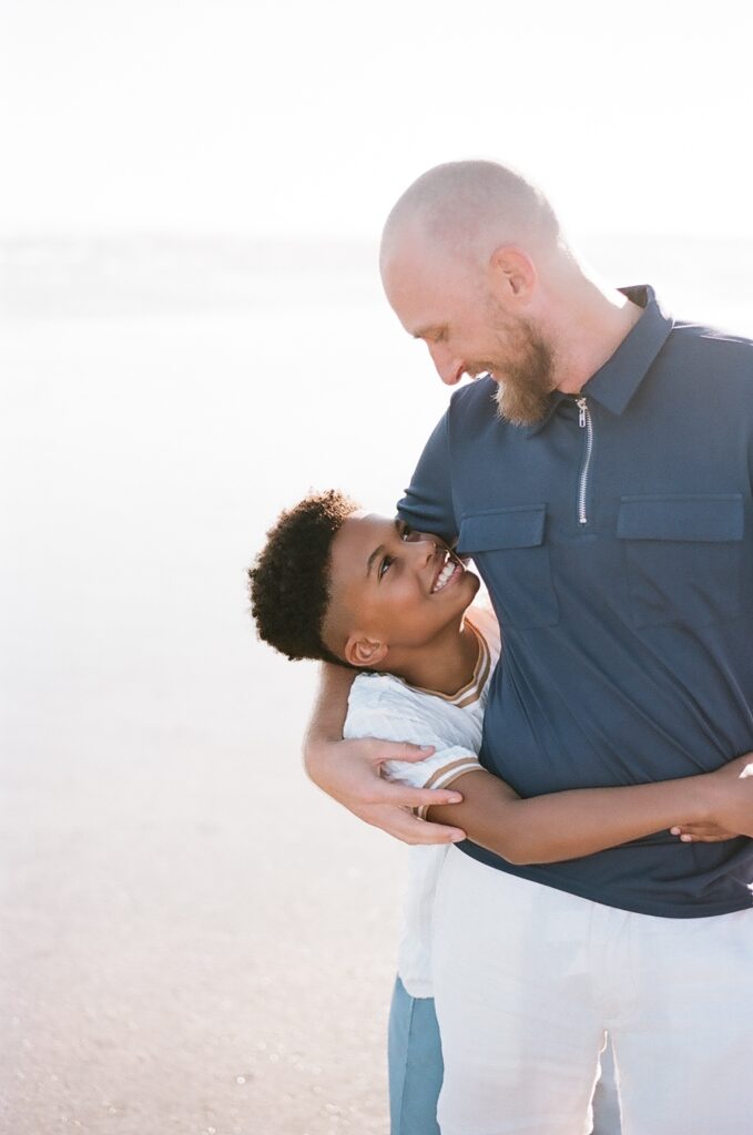 A heartwarming moment captured on Isle of Palms beach, featuring a white father embracing his black son with wide smiles. This touching image beautifully represents the bond between a father and his son, celebrating love. Photographed by Charleston photographer Kelsey Halm, this photo captures the joy and connection within a diverse family.