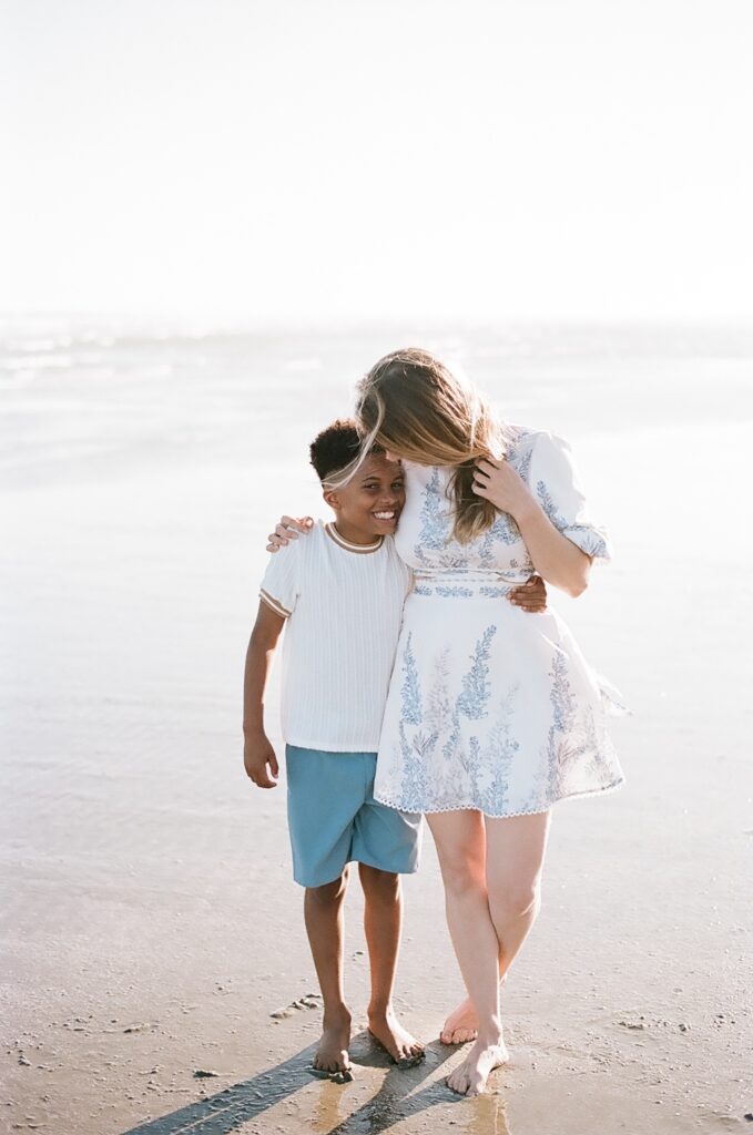 A beautiful film portrait of a mother and her son standing together on the Isle of Palms beach, both smiling at each other. The mother wears a white sundress, while her son wears a blue shirt and shorts. The soft sunlight of the beach enhances the warmth of their skin and brings out the love and bond between them.