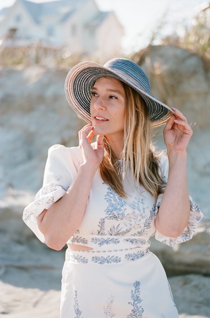 A captivating film photo of a woman on Isle of Palms beach, looking away from the camera with a sense of tranquility. She enhances her beach ensemble with a fashionable blue sun hat that adds a touch of elegance. Her attire consists of a beautiful white sundress adorned with delicate blue florals, exuding a sense of grace and style.