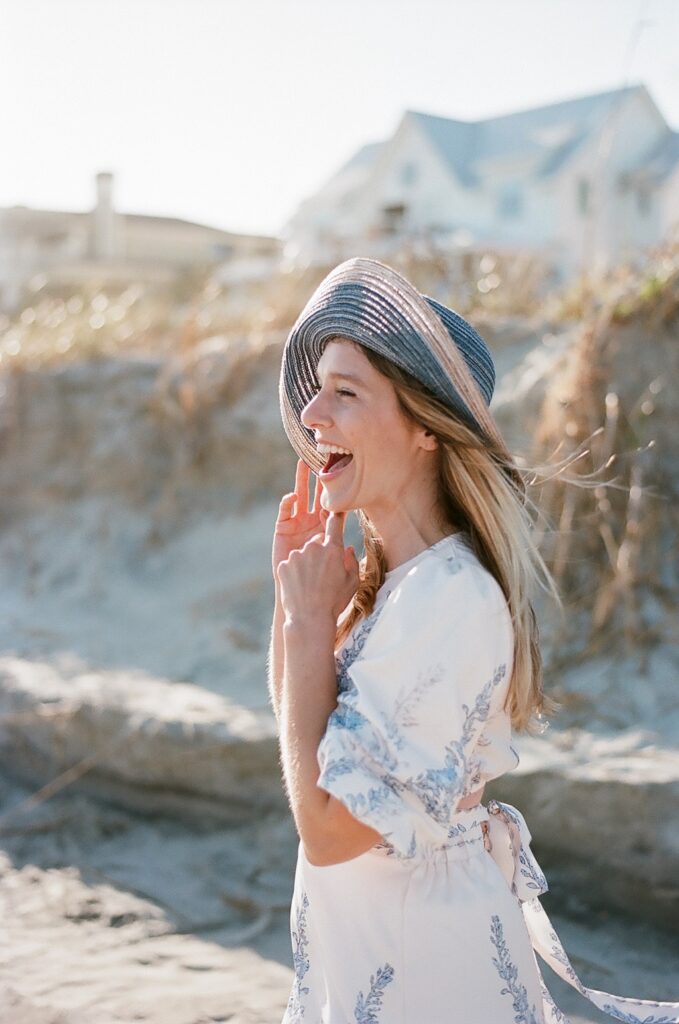 A captivating film photo capturing a joyful moment of a woman on Isle of Palms beach. She looks away from the camera, filled with genuine laughter and happiness. Her blue sun hat sits gracefully atop her head, adding a charming touch to her attire. She wears a lovely white sundress adorned with elegant blue florals, creating a beautiful contrast against the sandy backdrop.