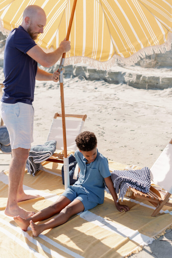 A captivating film photograph showcasing a father setting up a vibrant yellow and white fringed beach umbrella on a cheerful yellow and white beach blanket. His curious son sits nearby. The scene is adorned with two comfortable white slingback chairs and two stylish navy striped beach towels, creating a perfect beach setup. This delightful moment captured on Isle of Palms beach is photographed by Charleston photographer Kelsey Halm.