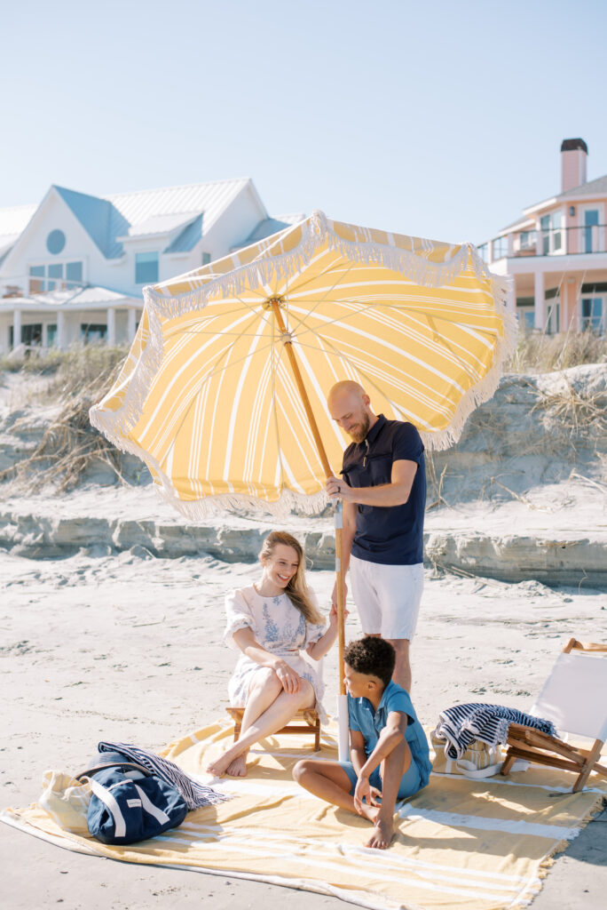 A film photograph capturing a loving family on Isle of Palms beach. The father is setting up a cheerful yellow and white fringed beach umbrella on a vibrant yellow and white beach blanket, while holding his wife's hand. Their adorable son sits in front of them on the blanket, filled with excitement. The scene is enhanced by the presence of two comfortable white slingback chairs and two stylish navy striped beach towels. This beautiful moment, captured by Charleston photographer Kelsey Halm, radiates joy and togetherness.