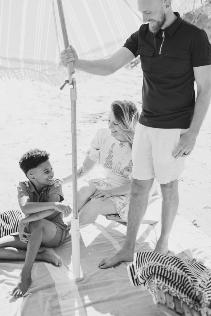 A heartwarming black and white image of a joyful family of three gathered under a beach umbrella, sharing smiles and happiness with one another.