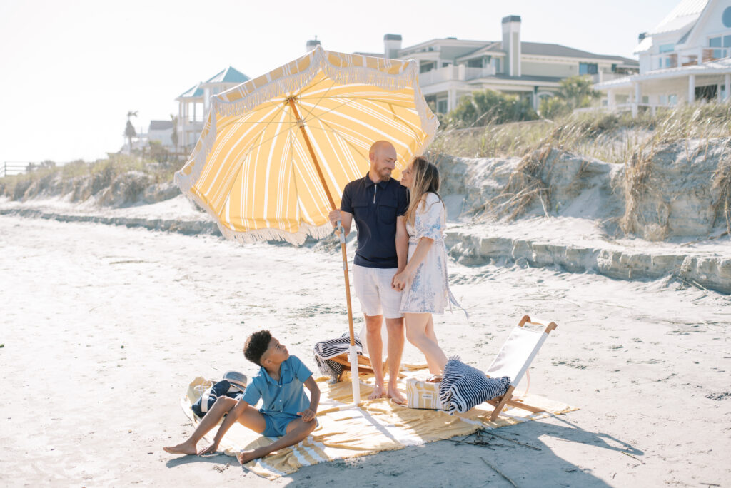 A beautiful moment captured on Isle of Palms beach by Charleston vacation photographer Kelsey Halm. The image features a loving husband and wife standing under a yellow and white fringed beach umbrella on a cozy yellow and white beach blanket. They hold hands affectionately, while their son sits in front of them, observing their tender connection. Two white slingback chairs and two navy striped beach towels complete the inviting scene.