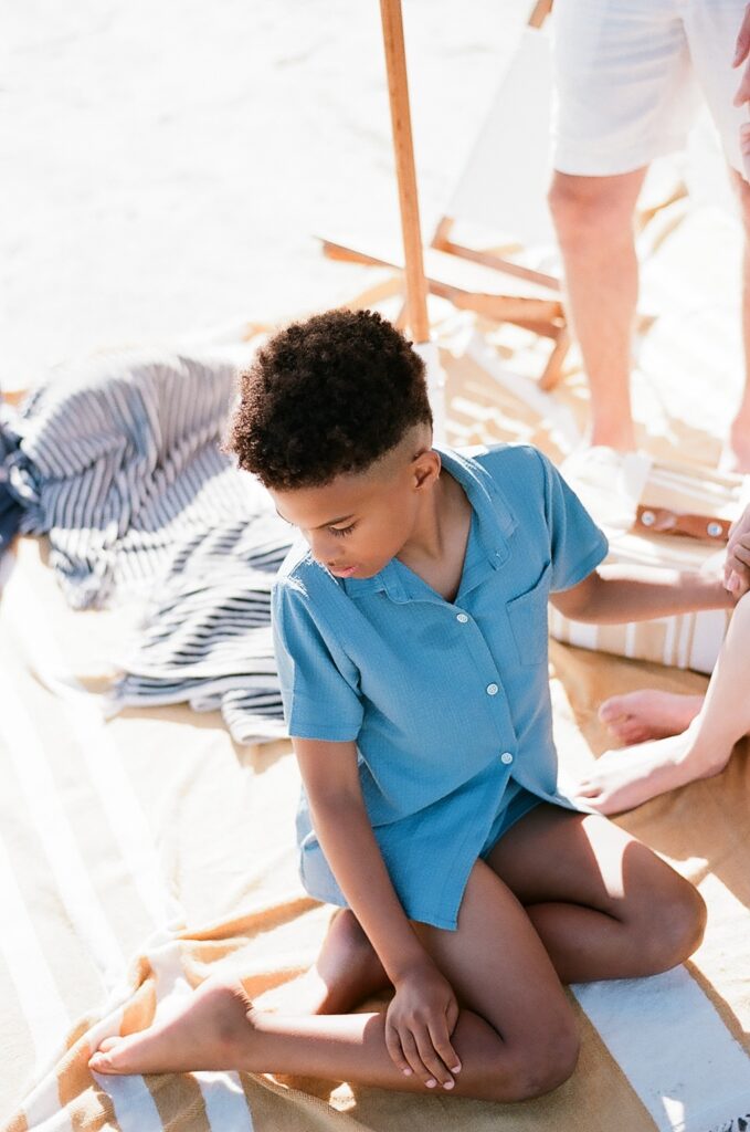 A captivating film photograph capturing an eight-year-old boy dressed in a stylish blue shirt and shorts ensemble. He sits on a cozy yellow and white beach blanket, holding his mom's hand while glancing down at his feet. Nearby, a stack of navy striped towels adds a touch of seaside charm to the scene.