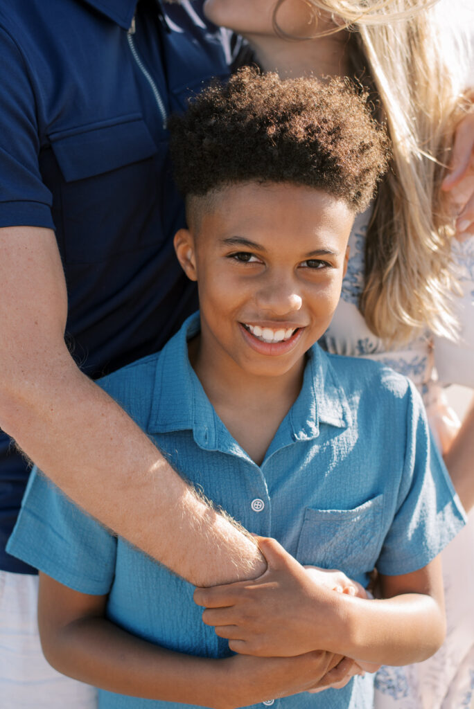 A heartwarming close-up of a cheerful young boy leaning back against his parents, grasping his father's arm affectionately. The parents, barely visible in the background, share an intimate moment with their noses touching. The son's radiant smile captivates as he gazes directly at the camera. This touching family portrait captures the bond and happiness shared between parents and child.