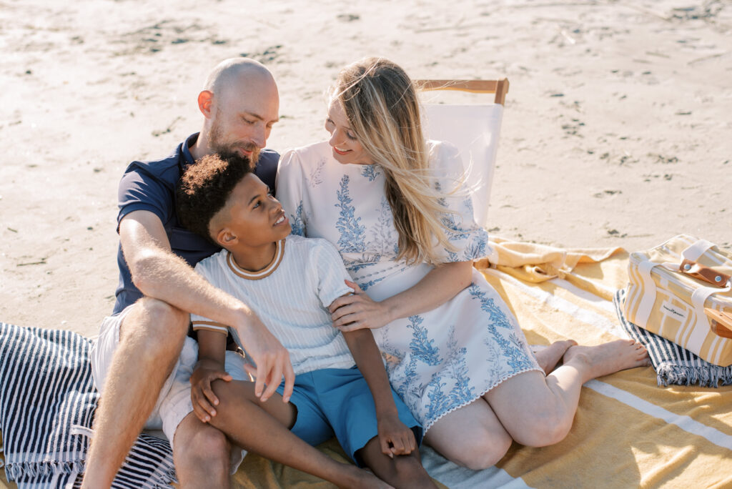 A happy family of three enjoying quality time together on a yellow beach blanket at Isle of Palms beach. Their genuine smiles and loving expressions capture the joy of the moment. In the background, two navy striped towels and a yellow and white beach bag complete the scene.