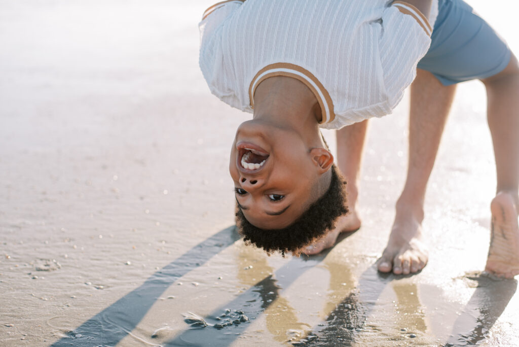 A playful young boy flips over backwards on the sandy beach, giggling with joy, while securely holding his father's hands. The father, standing strong and supportive, shares in his son's excitement. This lighthearted moment captures the fun and connection between father and son on a memorable beach outing.