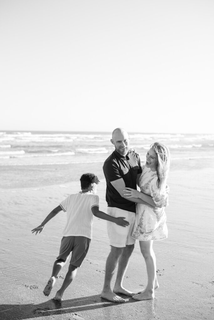  joyful black and white photograph of a loving family of three at Isle of Palms beach. The husband and wife share a warm embrace, their laughter filling the air, as their playful son runs in circles around them. The candid moment captures the essence of family happiness and togetherness.
