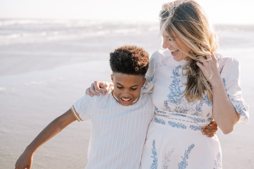 A loving mother and son share a tender moment on Isle of Palms beach, their arms wrapped around each other's backs as they burst into laughter. This heartwarming scene, captured by Charleston photographer Kelsey Halm, showcases the deep bond and joy between a mother and her child. The natural beauty of the beach serves as a stunning backdrop, adding to the charm of this precious moment. Ideal for family photography and cherished memories.