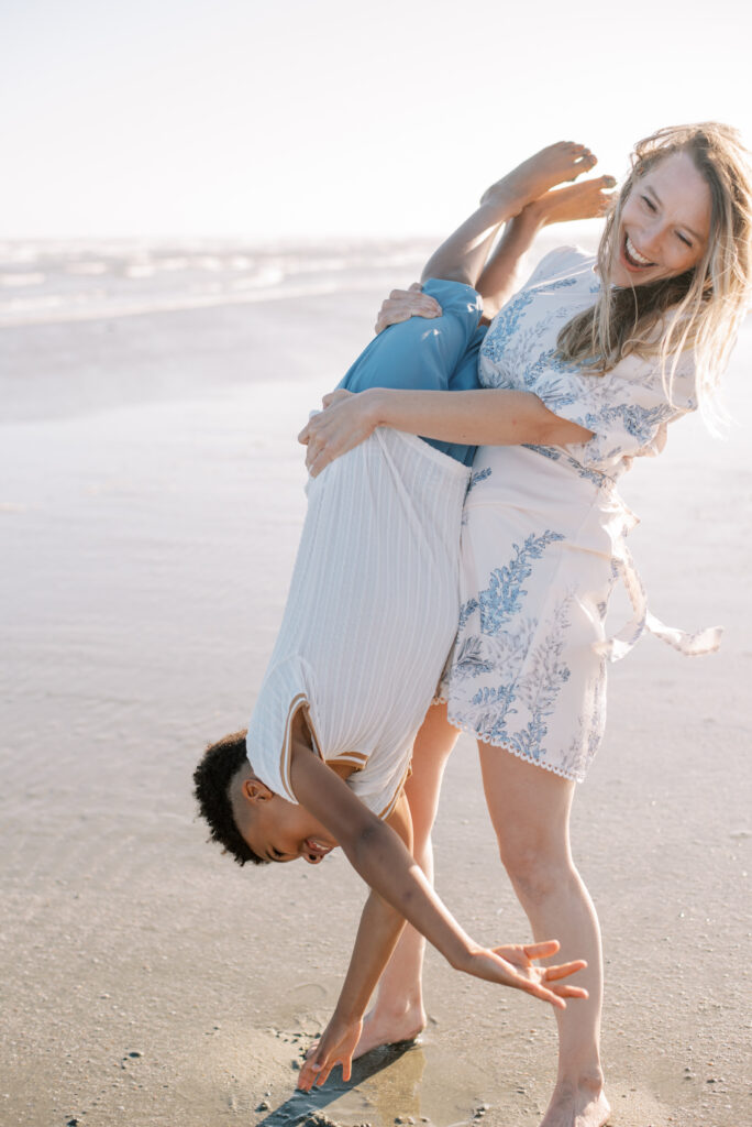 A playful mother holds her joyful eight-year-old son upside down on Isle of Palms beach, both laughing as he reaches out to touch the soft sand. This delightful moment captures the love and laughter shared between a mother and her child. The beach provides a picturesque setting for this fun-filled interaction, creating a memorable experience. Photographed by Charleston photographer Kelsey Halm