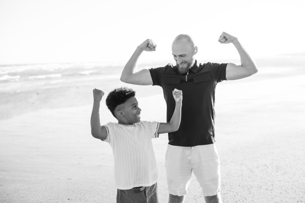 Black and white image of A father and son stand side by side on a sandy beach, striking playful muscle poses as they compare their strength, sharing hearty laughter. This heartwarming moment captures the bond between a father and his son, filled with joy and camaraderie. The scenic backdrop of the beach adds to the charm of the image, providing a natural and relaxed atmosphere. This captivating photograph, depicting the special connection between a father and his child, is a perfect fit for family-oriented content and brings a sense of happiness and togetherness.