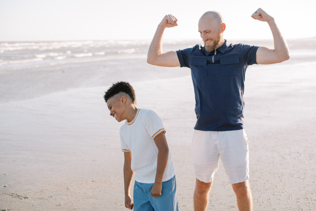 A father and son stand side by side on a sandy beach, striking playful muscle poses as they compare their strength, sharing hearty laughter. This heartwarming moment captures the bond between a father and his son, filled with joy and camaraderie. The scenic backdrop of the beach adds to the charm of the image, providing a natural and relaxed atmosphere. This captivating photograph, depicting the special connection between a father and his child, is a perfect fit for family-oriented content and brings a sense of happiness and togetherness.