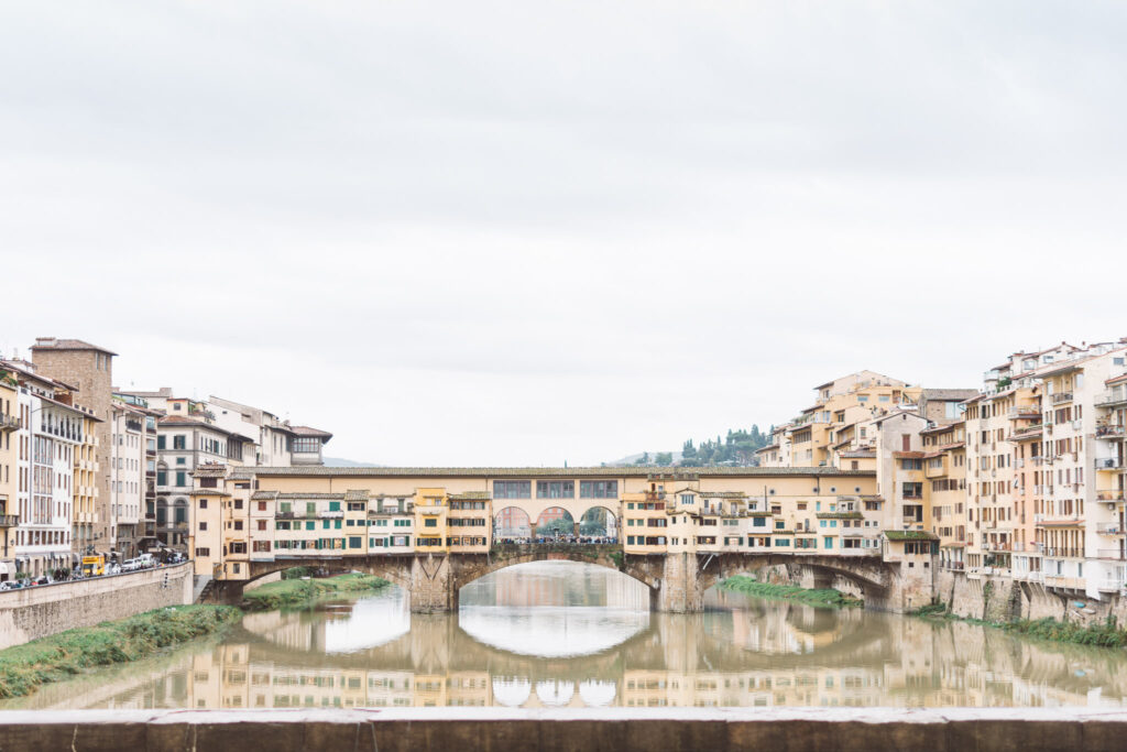 Scenic view of the Ponte Vecchio bridge, an iconic landmark of Florence, Italy, as seen from the Arno river. The historic bridge is lined with shops and has arched passageways, while the river flows underneath. In the background, there are buildings with terracotta roofs and lush greenery