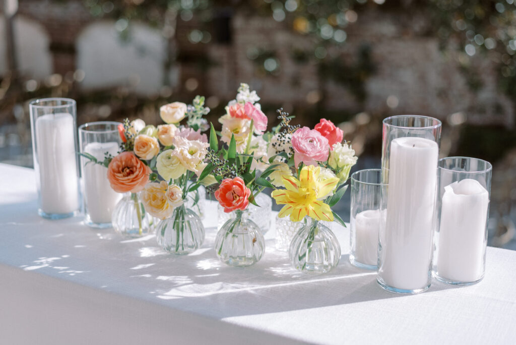 Colorful floral arrangement with pillar candles and small vases adorns a side table with white linens at a bridal brunch in the Gadsden House, Charleston. The beautiful flowers include a mix of pink, white, and yellow blossoms, creating a stunning visual display for the brunch event.