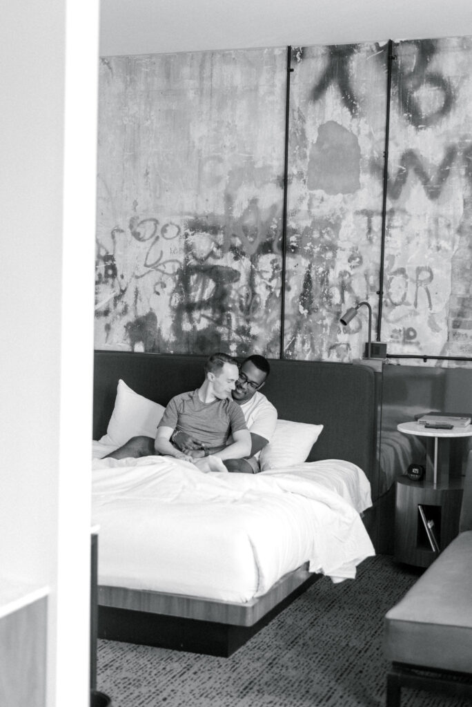 Black and white photograph of two engaged men sitting closely together on a hotel bed, sharing an intimate moment. They are dressed casually in comfortable clothing, and their faces are illuminated by the soft natural light coming from a nearby window. The image captures the tenderness and love between the couple in the quiet moments leading up to their wedding day. Photographed by Kelsey Halm