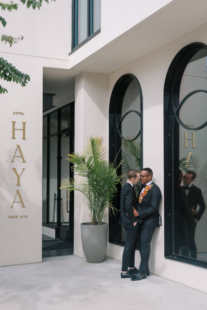 Two grooms wearing custom black tuxedos share a kiss in front of the white stucco walls and large black decorative windows of the historic Hotel Haya in Ybor City, Tampa
