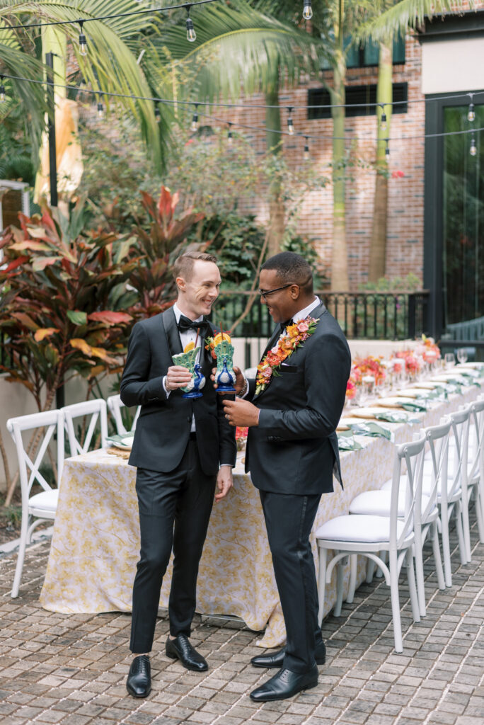 Two grooms are shown in the midst of a celebratory toast, holding unique peacock cocktail glasses. The background is the outdoor courtyard of the Hotel Haya in Ybor City, featuring a long wedding reception table lined with vibrant floral arrangements in shades of orange, pink, and yellow. The table is dressed in a gold gingko patterned linen with green napkins adorned with white florals, rattan chargers, white plates, and gold flatware with white handles.